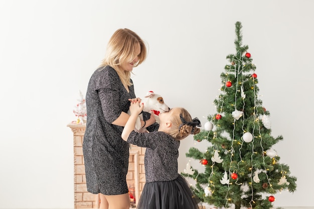 Férias e conceito do animal de estimação - mãe e filha brincando com o cachorro jack russell terrier na frente da árvore de natal em casa.