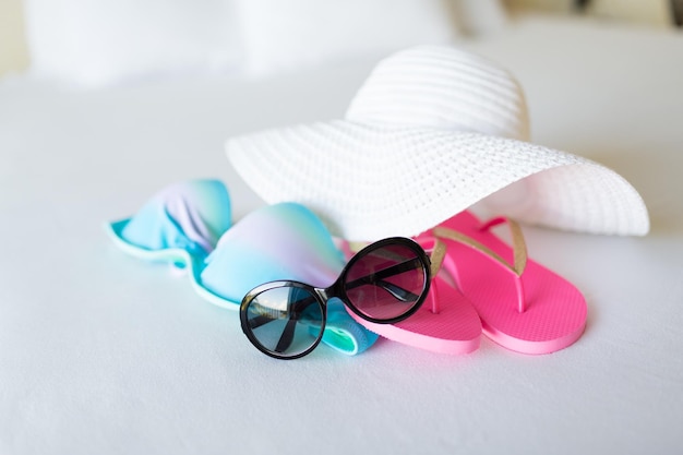férias, férias e conceito de viagem - close-up de top de biquíni, chapéu de palha, flip-flop e óculos de sol na cama do hotel