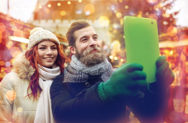 férias, inverno, natal, tecnologia e conceito de pessoas - casal feliz de turistas em roupas quentes tomando selfie com computador tablet pc andando na cidade velha