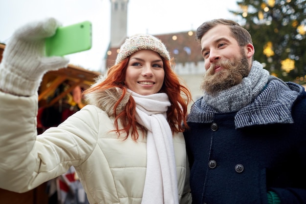 férias, inverno, natal, tecnologia e conceito de pessoas - casal feliz de turistas em roupas quentes tomando selfie com smartphone na cidade velha