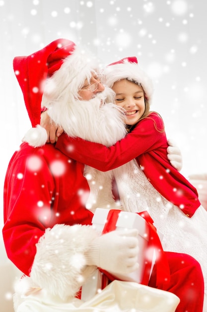 férias, natal, felicidade e conceito de pessoas - menina sorridente com caixa de presente abraçando papai noel em casa