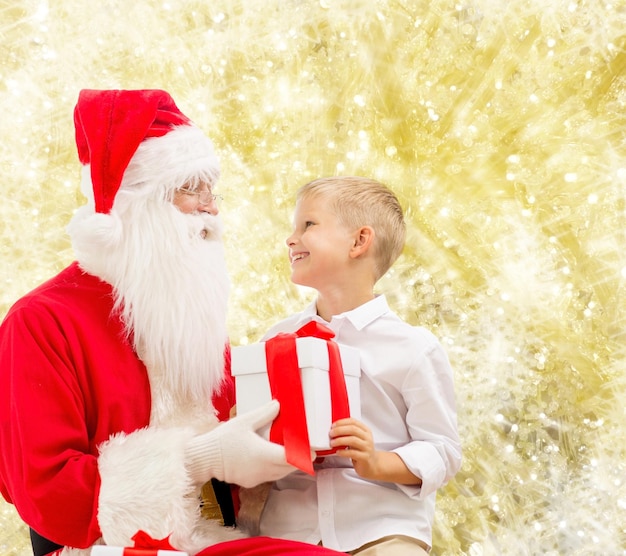 férias, natal, infância e conceito de pessoas - menino sorridente com papai noel e presentes sobre fundo de luzes amarelas