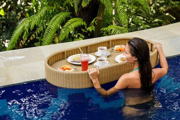 Férias no resort. jovem mulher feliz com um café da manhã flutuante na piscina.