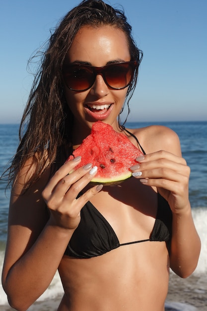 Férias, resort, conceito de turismo - férias de verão - jovem comendo melancia fresca na praia de areia. bela jovem come melancia na praia num dia quente de verão.