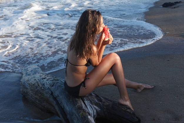 Férias, resort, conceito de turismo - férias de verão - jovem comendo melancia fresca na praia de areia. bela jovem come melancia na praia num dia quente de verão.