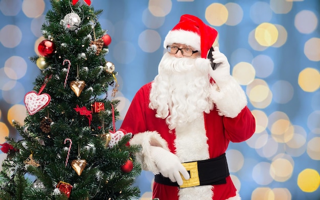 férias, tecnologia e conceito de pessoas - homem fantasiado de papai noel com smartphone e árvore de natal sobre fundo de luzes azuis