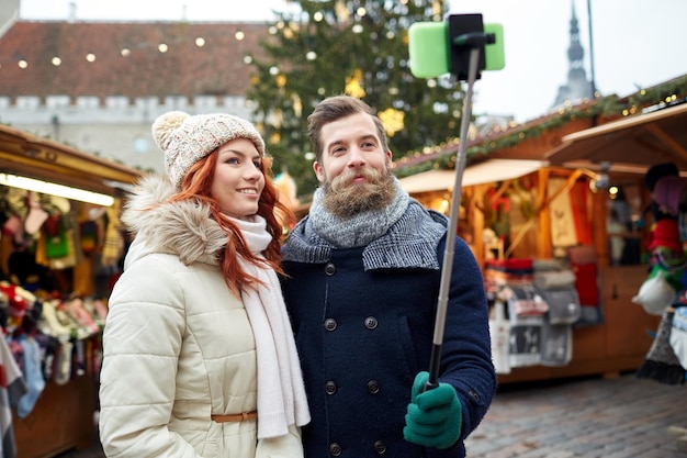 ferien, winter, weihnachten, technologie und personenkonzept - glückliches paar touristen in warmer kleidung, die mit smartphone-selfie-stick in der altstadt fotografieren