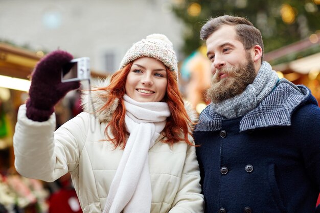 ferien, winter, weihnachten, technologie und personenkonzept - glückliches paar touristen in warmer kleidung, die selfie mit smartphone in der altstadt machen