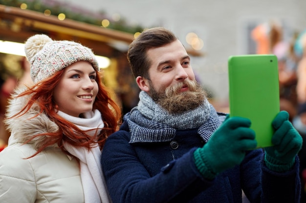 ferien, winter, weihnachten, technologie und personenkonzept - glückliches paar touristen in warmer kleidung, die selfie mit tablet-pc-computer machen, der in der altstadt spazieren geht