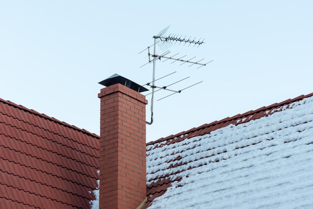 Fernsehantenne auf rotem Dach Fernsehantenne auf Dach mit blauem Himmel Ziegelsteinkamin und schneebedecktes Dach des Hauses