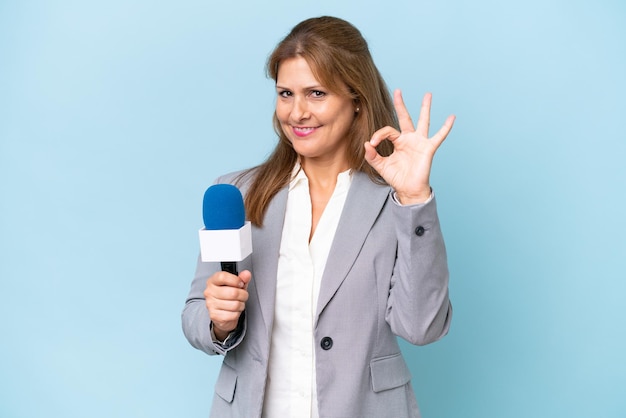 Fernsehmoderatorin mittleren Alters über isoliertem blauem Hintergrund, die mit den Fingern ein Ok-Zeichen zeigt