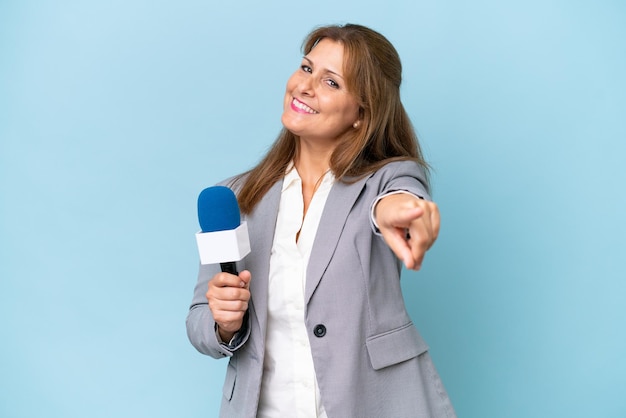 Fernsehmoderatorin mittleren Alters über isoliertem blauem Hintergrund, die mit fröhlichem Ausdruck nach vorne zeigt