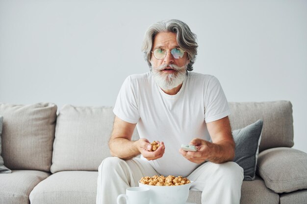 Fernsehsendung ansehen und Popcorn essen Älterer stilvoller moderner Mann mit grauem Haar und Bart drinnen