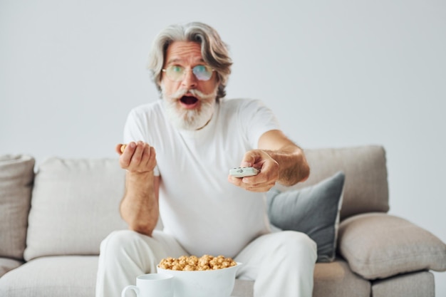 Fernsehsendung ansehen und Popcorn essen Älterer stilvoller moderner Mann mit grauem Haar und Bart drinnen