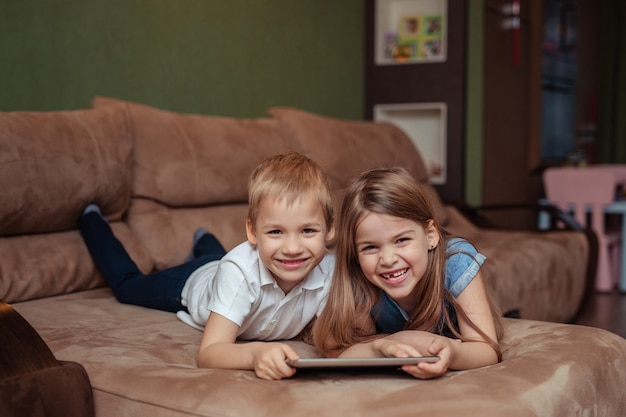 Fernunterricht zu Hause. Zwillinge Bruder und Schwester lernen zu Hause mit einem Tablet. Sie sind glücklich und lachen, während sie auf der Couch liegen