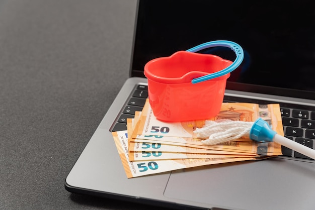 Ferramentas de limpeza em miniatura e euros em dinheiro no teclado do laptop Serviços de limpeza on-line Conceito de lavagem de dinheiro pela Internet