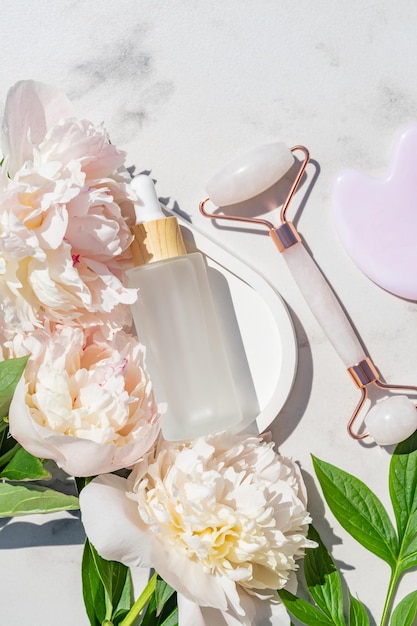 Ferramentas de massagem facial Gua Sha rosa Rolo de jade de quartzo rosa e óleo facial de beleza em fundo de mármore branco com peônias Tratamento anti-idade e tonificação em casa