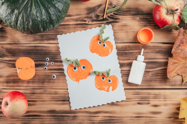 Fertige Halloween-Karte mit Kürbis-Apfel-Prints liegt neben Kleber und Farbe auf Holzoberfläche, Draufsicht