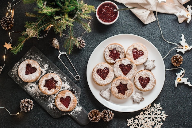 Fertige Linzer Kekse mit Beerenmarmelade auf einem Teller und Weihnachtsschmuck auf dunklem Hintergrund. Weihnachtsleckereien kochen. Lebensstil. Ansicht von oben