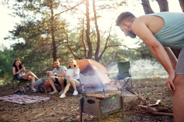 Festa, acampamento de grupo de homens e mulheres na floresta. eles relaxam, cantam uma música e cozinham churrasco na grama verde. as férias, verão, aventura, estilo de vida, conceito de piquenique