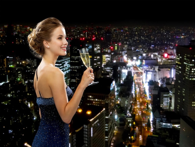 Festa, bebidas, feriados, luxo e conceito de celebração - mulher sorridente em vestido de noite com copo de vinho espumante sobre o fundo da cidade à noite