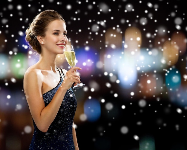 festa, bebidas, feriados, natal e conceito de pessoas - mulher sorridente em vestido de noite com copo de vinho espumante sobre luzes noturnas e fundo de neve