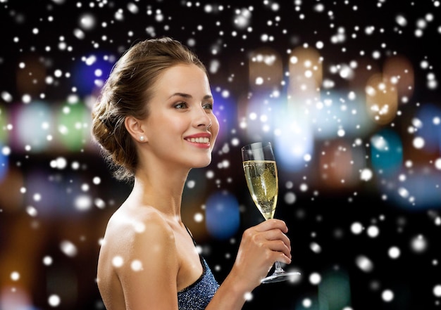 festa, bebidas, feriados, natal e conceito de pessoas - mulher sorridente em vestido de noite com copo de vinho espumante sobre luzes noturnas e fundo de neve