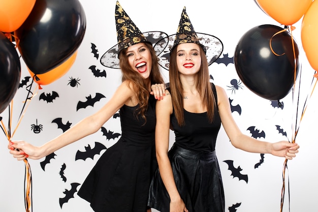 Festa de Halloween. Duas lindas mulheres em vestidos pretos e chapéus de bruxa seguram balões pretos e laranja no fundo da parede com morcegos. .
