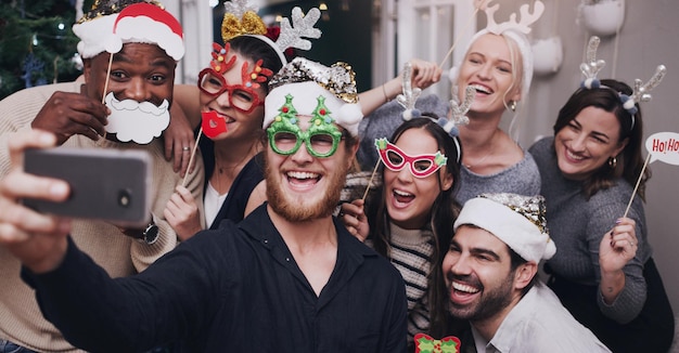 Festa de Natal e amigos tirando uma selfie em um telefone junto com adereços engraçados e bobos Diversidade pessoas festivas e felizes tirando fotos em um smartphone em um evento festivo de natal em uma casa