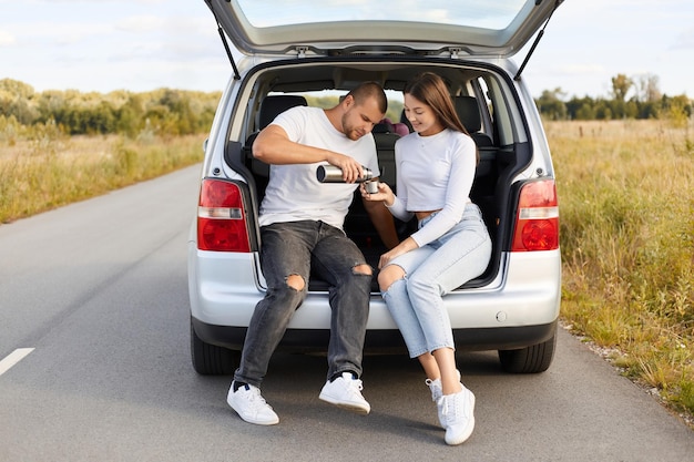 Festa do chá no porta-malas do carro Retrato de casal apaixonado bebe chá quente da garrafa térmica sentado no porta-malas do carro família vestindo camisas brancas e jeans viajando juntos pare para descansar e haning lanche