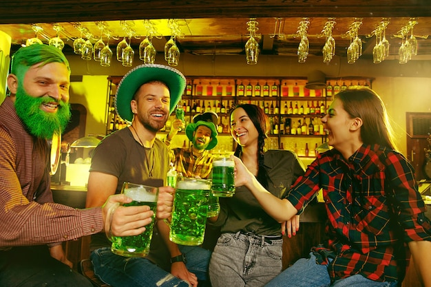 Festa do dia de são patrício. amigos felizes estão comemorando e bebendo cerveja verde. homens e mulheres jovens usando chapéus verdes. pub interior.