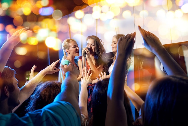 festa, feriados, celebração, vida noturna e conceito de pessoas - mulheres jovens felizes cantando karaokê na boate atrás da multidão de fãs de música