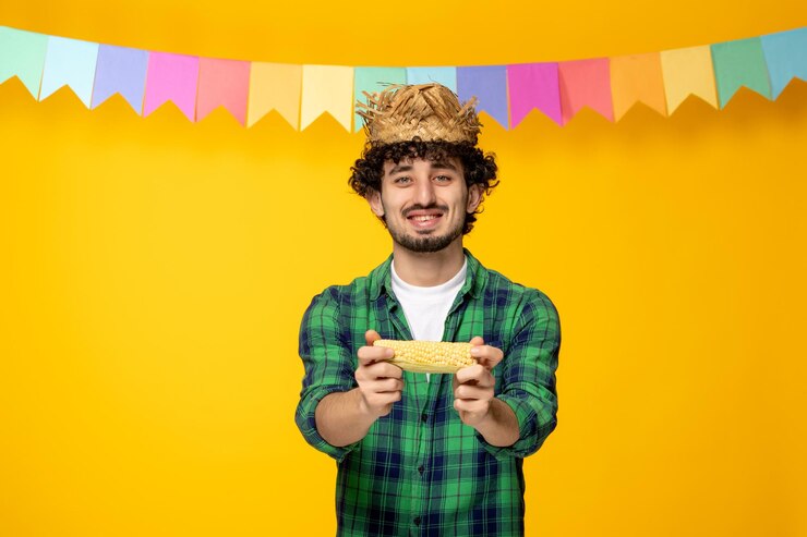 Foto festa junina jovem bonitinho de chapéu de palha e bandeiras coloridas festival brasileiro dando milho