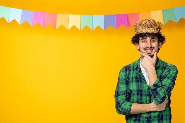 Foto festa junina jovem bonitinho de chapéu de palha e bandeiras coloridas festival brasileiro segurando o queixo sorrindo