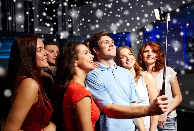 festa, tecnologia, vida noturna e conceito de pessoas - amigos sorridentes com smartphone e monopé tomando selfie no clube e efeito de neve