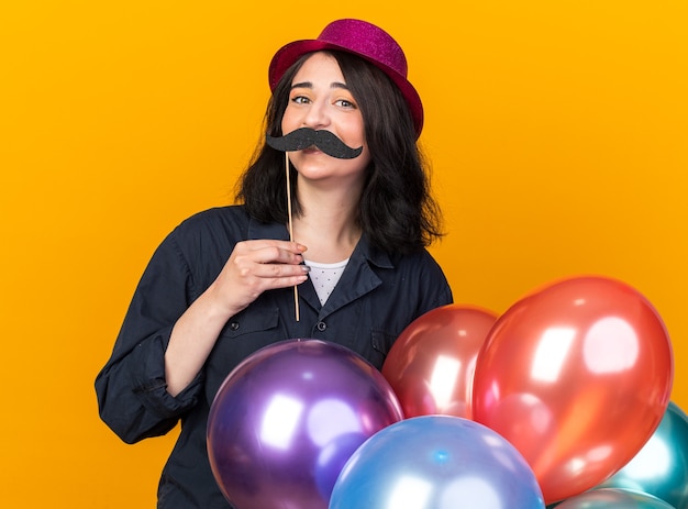 Festeira jovem caucasiana engraçada usando chapéu de festa segurando um monte de balões nas costas e bigode falso em um palito acima dos lábios isolados na parede laranja