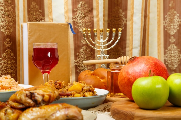 Festlich gedeckter Tisch für eine Mahlzeit an Rosh Hashanah, Challah, Honig, Granatapfel, Äpfel, Wein und traditionelle Gerichte neben Menorah und Siddur