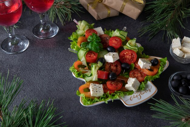 Festliche Häppchen mit Gurken, Tomaten und Käse auf dem Teller als Weihnachtsbaum serviert, auf dunkelgrauem Hintergrund mit zwei Gläsern Wein.