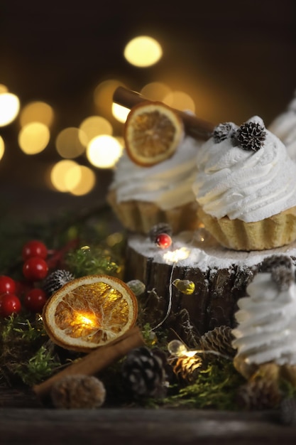 Festliche Kuchen in Form von Törtchen mit Sahne auf dunklem Hintergrund mit Moos, Zimt, Orangenkegeln. Garland ist um Törtchen, Weihnachtsstimmung, festliche Tafel