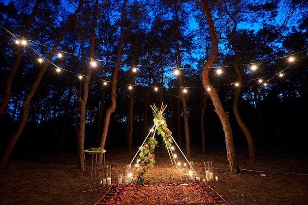 Festliche Lichterkettenbeleuchtung auf Boho-Tipi-Bogendekor auf Hochzeitszeremonie im Freien in Kiefer