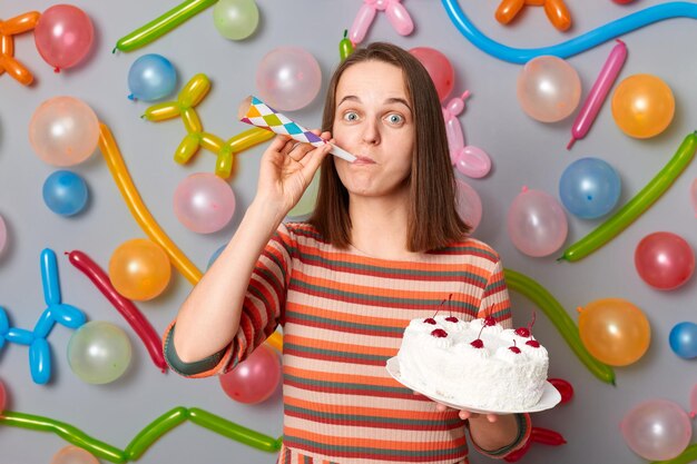 Festliche, lustige, glückliche Frau mit braunen Haaren, die ein gestreiftes Kleid trägt und einen Kuchen hält, der ein Partyhorn bläst, das Spaß auf einer Party hat, die an einer grauen Wand steht, die mit bunten Luftballons dekoriert ist