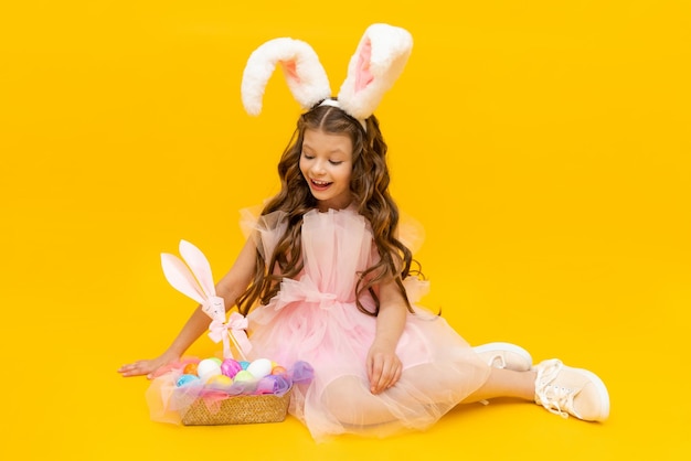 Festliche Ostern Ein kleines Mädchen mit Hasenohren und einem Korb mit bunten Eiern lächelt breit auf einem gelb isolierten Hintergrund