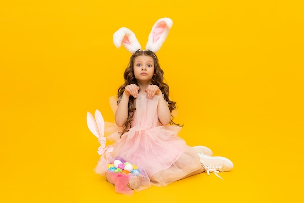 Festliche Ostern Ein kleines Mädchen stellt einen Osterhasen mit einem Korb voller bunter Eier dar. Ein Kind mit Hasenohren auf einem gelb isolierten Hintergrund