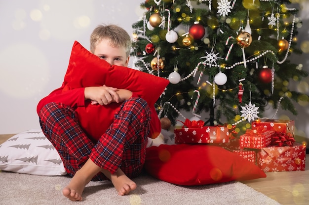 Festlicher lustiger Junge im Weihnachtspyjama und mit gemütlichem Kissen sitzt in der Nähe des Weihnachtsbaums mit Geschenken