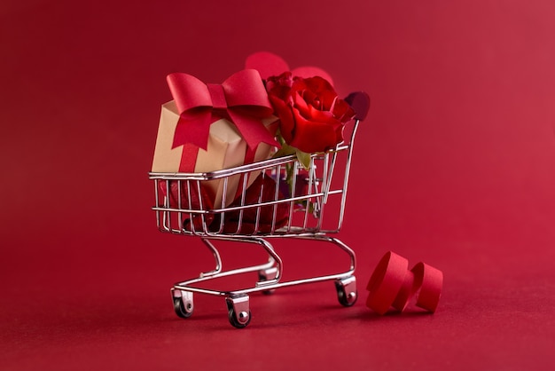 Festliches Verkaufskonzept des Valentinstags mit Geschenkboxrose und roten Papierherzen im Einkaufswagen gegen ein Rot.