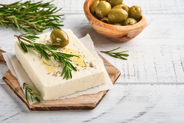 Feta-Käse-Würfel mit Rosmarin, Oliven und Olivenöl-Sauce in weißer Schüssel auf hellgrauem Hintergrund. Traditioneller griechischer hausgemachter Käse. Selektiver Fokus.