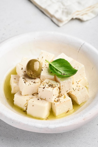 Feta-Käse-Würfel mit Rosmarin, Oliven und Olivenöl-Sauce in weißer Schüssel auf hellgrauem Hintergrund. Traditioneller griechischer hausgemachter Käse. Selektiver Fokus.