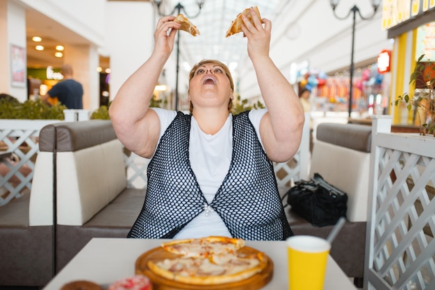 Fette Frau, die Pizza im Einkaufszentrumrestaurant isst, ungesundes Essen. Übergewichtige weibliche Person am Tisch mit Junk-Dinner, Fettleibigkeitsproblem