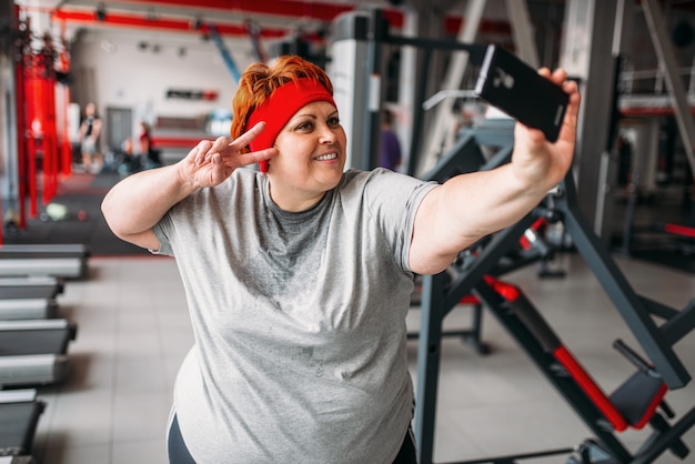 Fette verschwitzte Frau macht Selfie gegen Trainingsgeräte im Fitnessstudio. Kalorien brennende, fettleibige weibliche Person im Sportverein