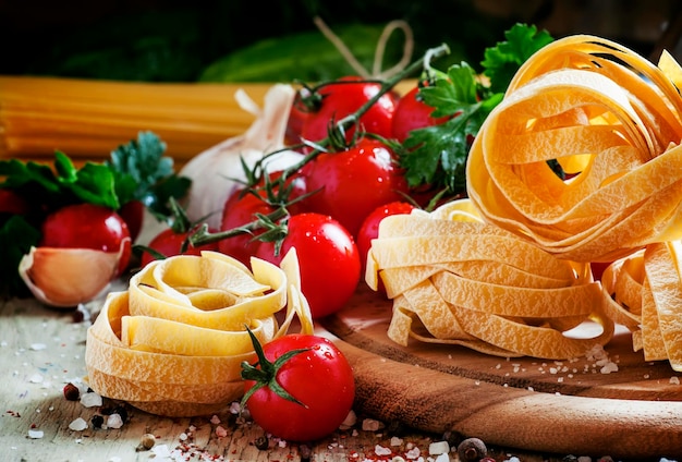 Fettuccine de pasta italiana seca en forma de nidos tomates cherry perejil eneldo ajo aceite de oliva en una jarra sal y especias en el antiguo fondo de madera enfoque selectivo